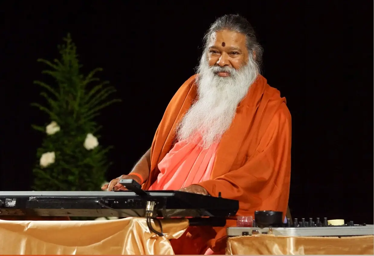 Sri Ganapathy Sachchidananda Swamiji at the synthesiser in saffron robe, in a serene evening setting.